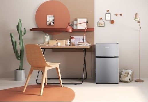 A small silver mini fridge beside a desk, chair, and floor lamp