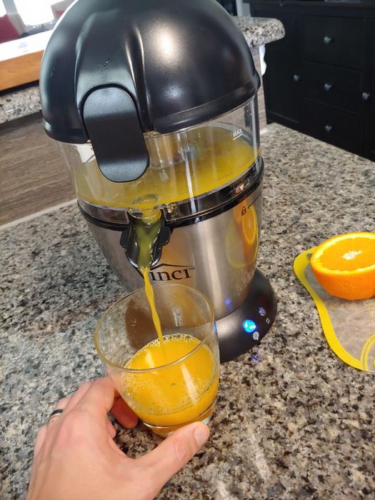 A juicer on the kitchen counter pouring fresh orange juice into a glass, with sliced oranges placed nearby