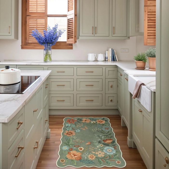 A contemporary kitchen featuring light wood cabinets, marble countertops, and a stovetop with a white pot. A floral rug and a vase of flowers on the counter enhance the decor