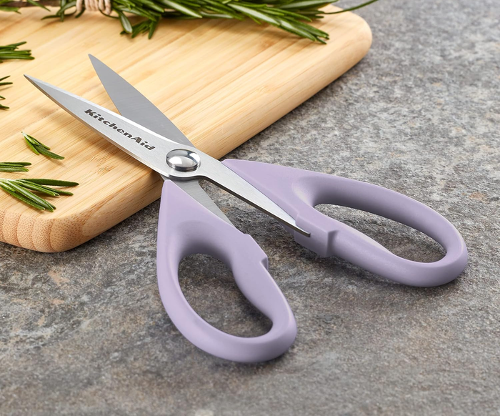 KitchenAid kitchen scissors with lavender handles resting on a counter beside a wooden chopping board and sprigs of rosemary
