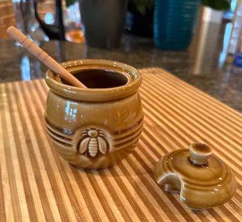 Ceramic honey pot featuring a bee design, wooden honey dipper inside, lid resting on a striped surface