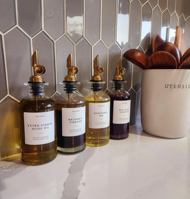 Four glass bottles of oil and vinegar, labeled Extra Virgin Olive Oil, Balsamic Vinegar, Vegetable Oil, and Red Wine Vinegar, positioned on a kitchen counter next to a utensil holder