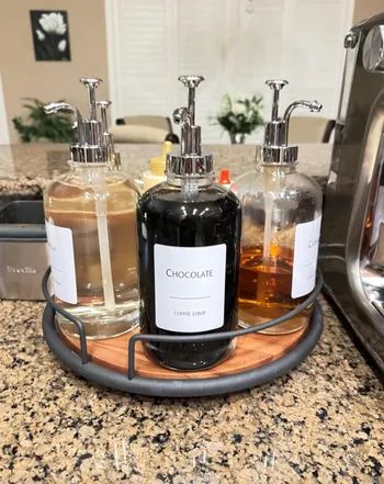 Three coffee syrup bottles labeled Chocolate, Caramel, and Vanilla, resting on a round tray with silver pump dispensers