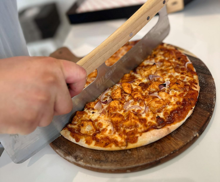 A person cutting a pizza topped with chicken and onions using a large knife on a wooden cutting board