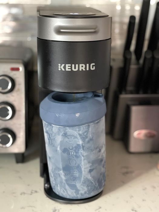a Keurig coffee maker brewing into a large, blue iced coffee tumbler resting on a kitchen counter