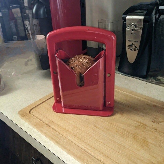 A loaf of bread positioned in a red slicer on a kitchen counter, with a chopping board and coffee maker in the background