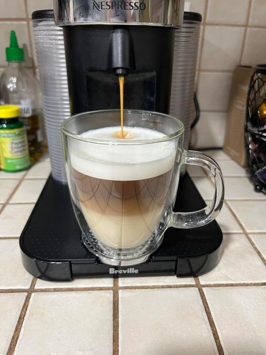 The Nespresso machine preparing a cup of coffee into a clear glass mug on a tiled countertop, with a rich espresso layer topped with a smooth frothy layer