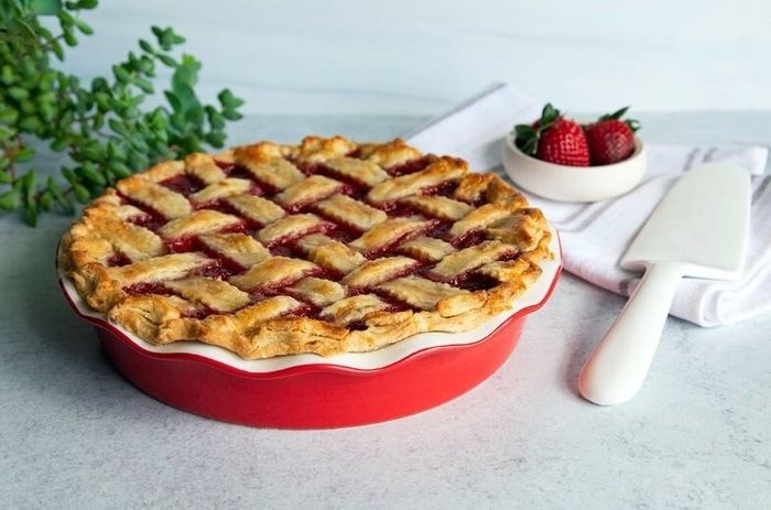 A lattice-topped pie in a bright red dish with fresh strawberries on the side, resting on a white napkin with a pie server