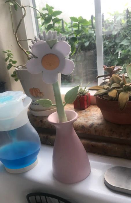 Flower-shaped dish brush in a pink vase on a windowsill, surrounded by various potted plants