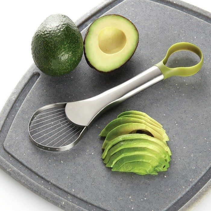 An avocado slicer next to a sliced avocado and avocado pieces on a cutting board, demonstrating its functionality for a Shopping article