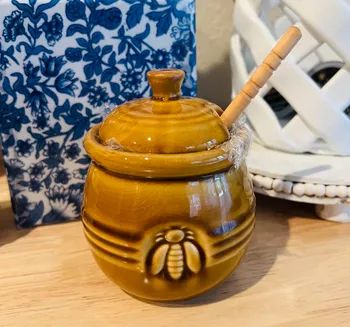 Ceramic honey pot with a lid and dipper, adorned with a bee motif, placed on a wooden surface next to decorative plates