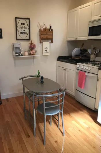 A small kitchen featuring a round table, two chairs, a stove, and a microwave. A wall art sign is visible, reading...