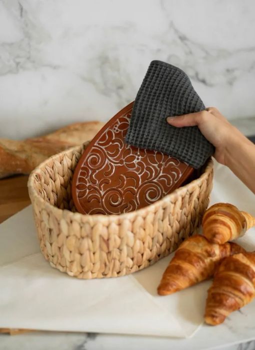 A hand carefully placing a decorative clay bread warmer into a woven basket with fresh croissants resting nearby.