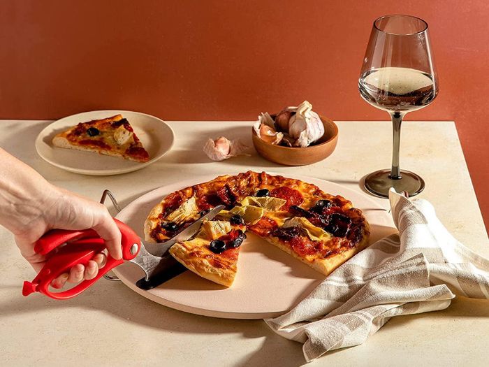 A person cutting a slice of pizza with red pizza scissors