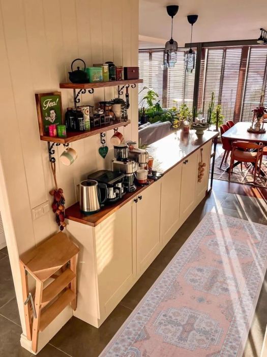 A cozy kitchen with coffee and tea items on wall shelves, a wooden countertop, and a sunlit dining area filled with plants in the background