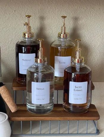 Four glass bottles of coffee syrup, each labeled Hazelnut, Toffee, Vanilla, and Salted Caramel, arranged on a wire rack with elegant gold pump tops