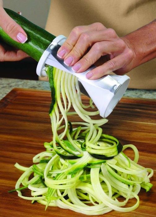 A hand turning the spiralizer, resembling a large pencil sharpener, to create zucchini noodles.