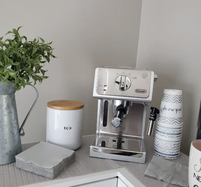 An espresso machine sitting on a countertop, surrounded by a plant, a tea canister, and neatly arranged stacked cups.