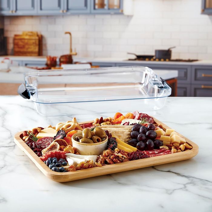 A food tray piled high with assorted cheeses, meats, olives, grapes, blueberries, nuts, and crackers resting on a kitchen counter, with a clear lid in the background.