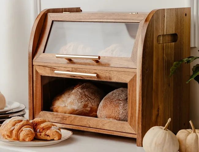 Wooden bread box with transparent glass doors showcasing bread loaves and croissants, complemented by neatly stacked plates and decorative white pumpkins nearby