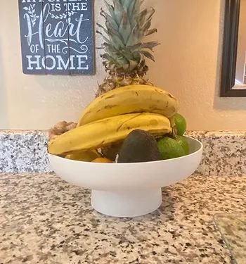 A petite white fruit bowl perched on a stand, filled with bananas and apples.