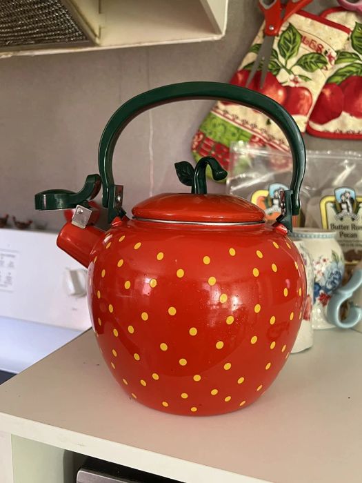 Strawberry-themed teapot with a green handle and yellow dots, sitting on a kitchen counter. The background features kitchen essentials and tea packages