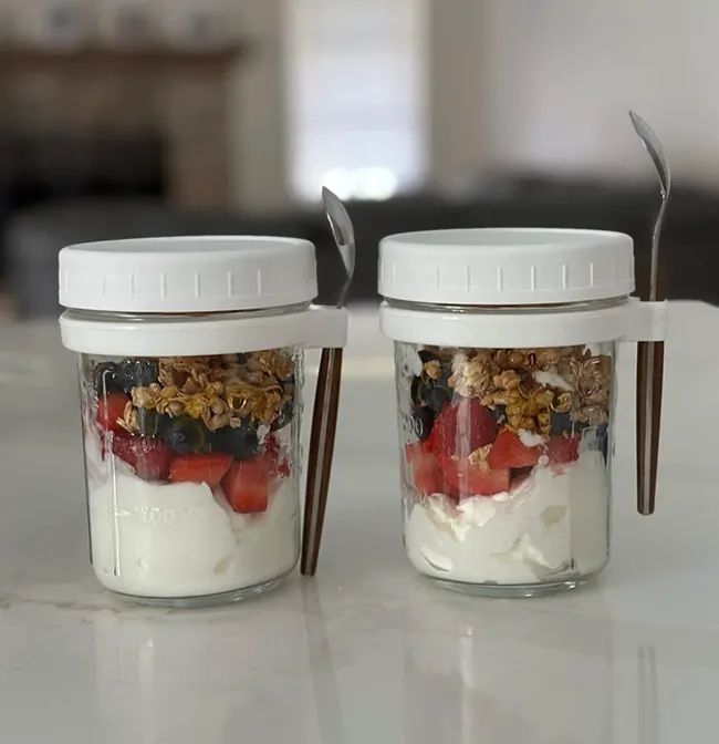 two small cylindrical jars filled with yogurt and granola, each with a twist-off white lid and tiny spoons attached