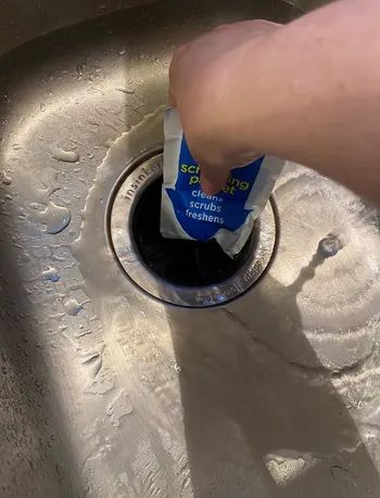A hand pouring Scrubbing Powder into a stainless steel sink drain