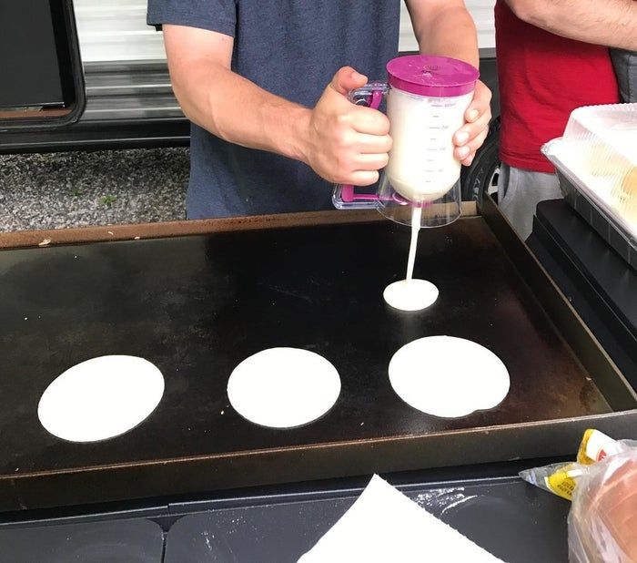 A person using a pancake dispenser to pour uniform circles of batter onto a hot griddle