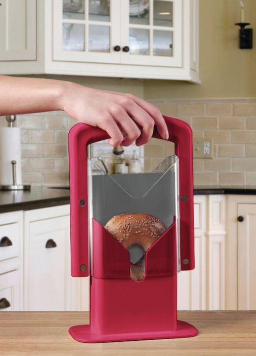 A hand is shown placing a bagel into a red countertop slicer in a modern kitchen with white cabinets in the background