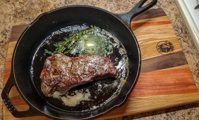 A sizzling steak cooking in a cast-iron skillet, topped with fresh herbs and a melting pat of butter