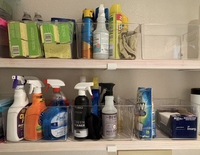 A neatly organized cleaning supply shelf with various bottles, sprays, sponges, and cleaning product boxes arranged neatly
