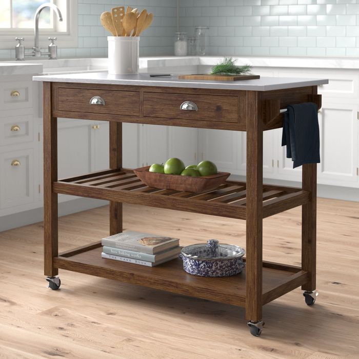 A wooden kitchen island on wheels featuring two drawers, two shelves, a towel rack, and a countertop with a bowl of fresh green apples and kitchen utensils on display