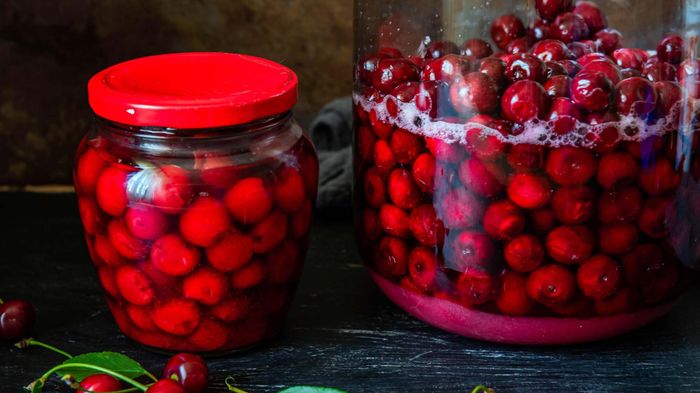 A jar of pickled cherries sits next to a larger container with cherries undergoing the pickling process.