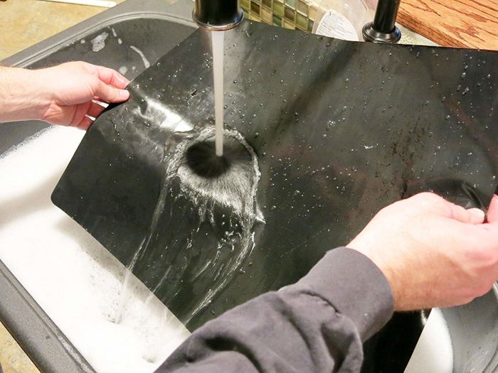 A person washing an oven liner under running water in the kitchen sink.