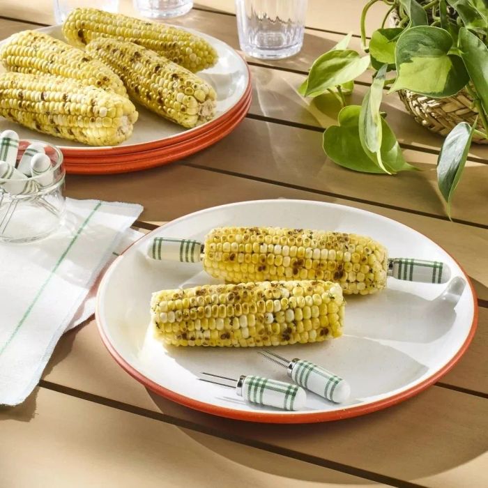 Plates of grilled corn on the cob with striped corn holders, placed on an outdoor wooden table alongside a cloth napkin, a jar, and a potted plant.