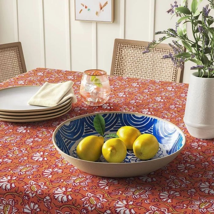 A bowl filled with four lemons sitting atop a patterned tablecloth, surrounded by plates, napkins, a drink, and a vase of flowers.