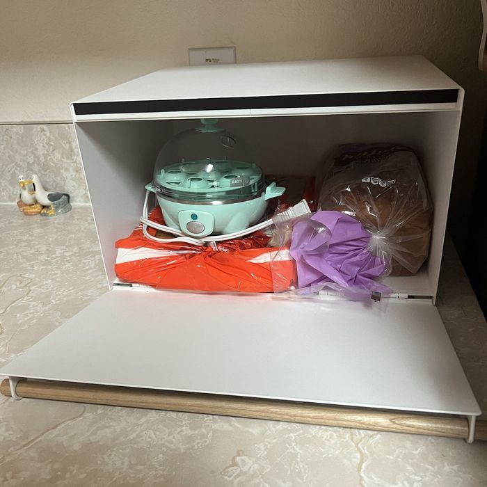 A bread box containing bread, an egg cooker, and a bag tucked behind a flap.