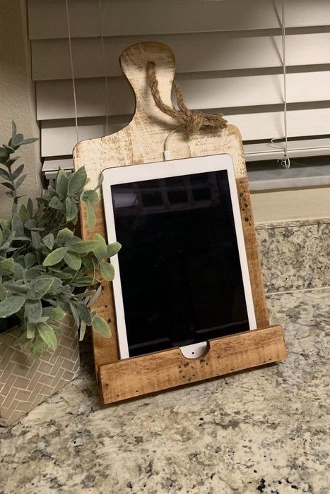 A wooden tablet holder stands proudly on a kitchen counter, accompanied by a leafy plant in a decorative pot, with fabric drapes and blinds softly filling the background.