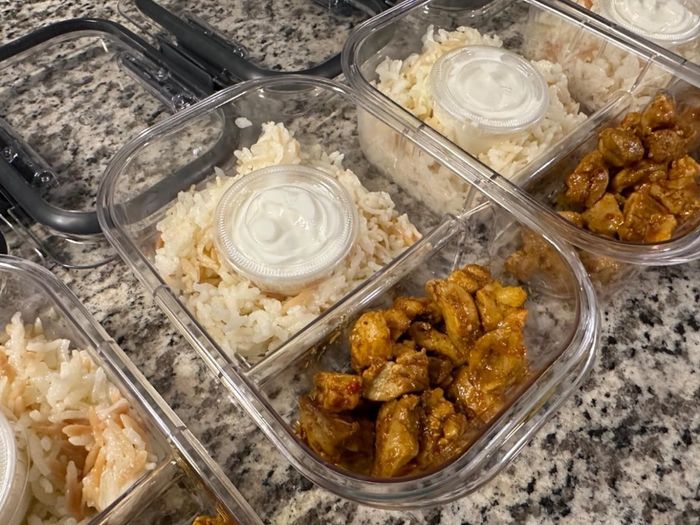 Meal prep containers filled with rice, pieces of chicken, and a lid covering some sauce.