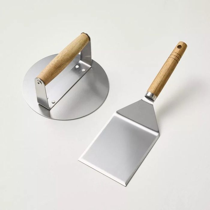 A metal griddle press and metal spatula with wooden handles, beautifully displayed against a white background. Featured in a shopping article.