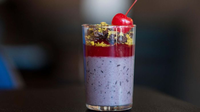 A single glass filled with a layered purple and red gelee, topped with pistachio powder, dried cherries, and a maraschino cherry.