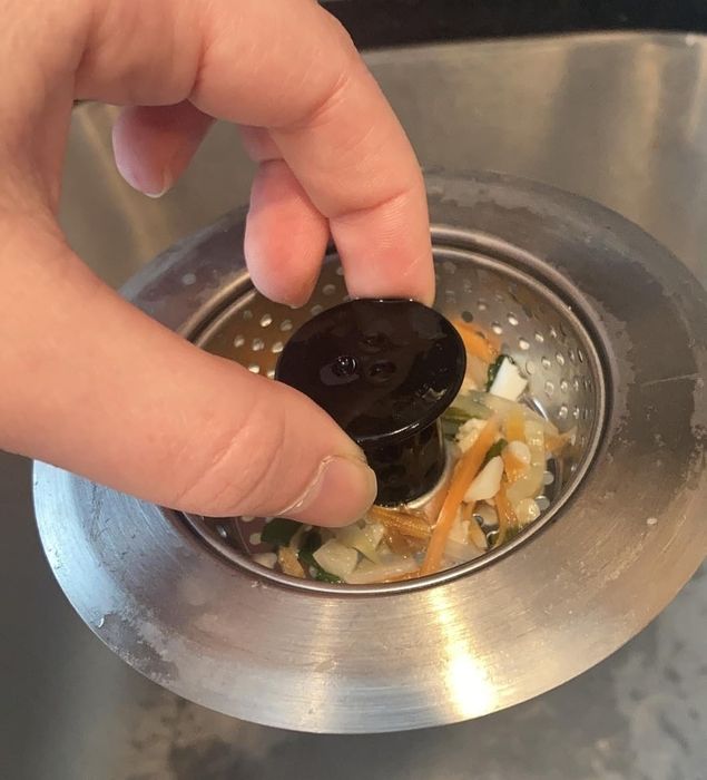 A close-up shot of a reviewer’s hand lifting a sink strainer full of food scraps from a shiny stainless steel sink, with soap bottles visible in the background.