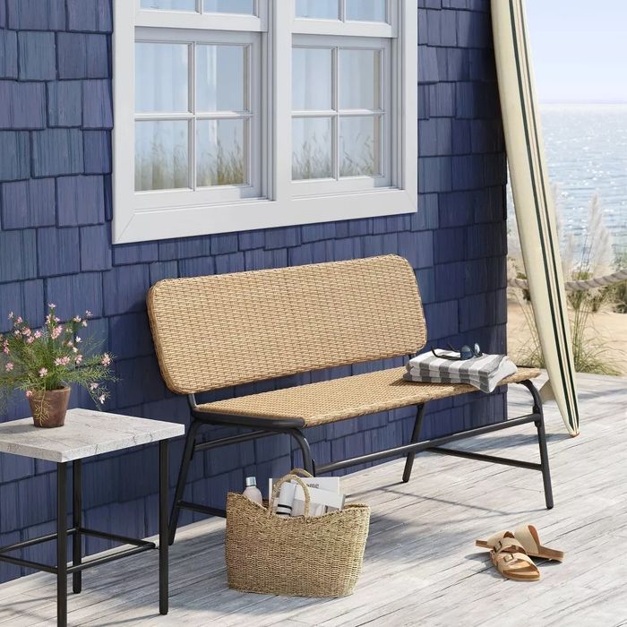 A patio setting featuring a wicker bench, side table, straw tote, and sandals near a blue beachside house