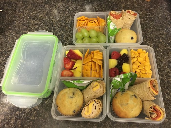 Four neatly organized meal prep containers filled with wraps, grapes, muffins, crackers, fruits, and cheese snacks