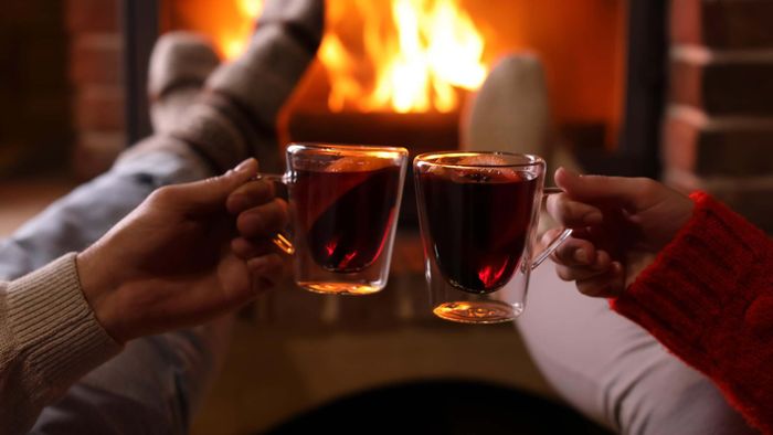 Two hands toasting with cups of mulled wine in front of a roaring fireplace.