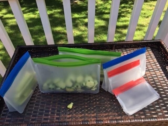 Reusable silicone storage bags arranged on a wicker tray, some filled with chopped vegetables. The bags come in various sizes and feature colorful zip closures