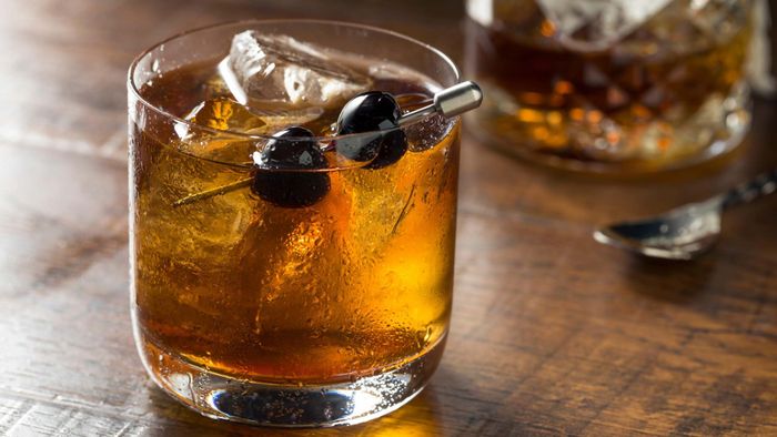 A rocks glass filled with ice and brown liquor, garnished with two brandied cherries on a metal skewer.