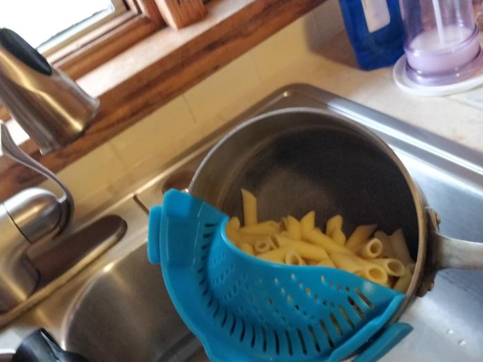 A metal pot of pasta being strained with a blue silicone colander over a kitchen sink