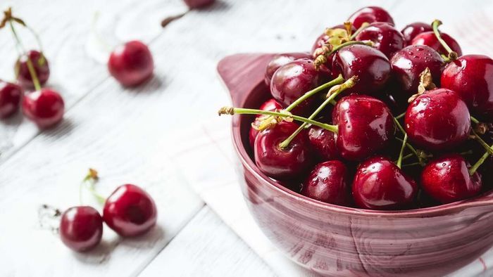 A red ceramic bowl filled with fresh cherries, surrounded by a scattering of loose cherries on the table.